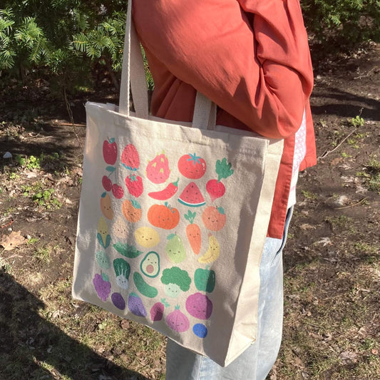 Tote bag with print of colorful fruits and vegetables with smiling faces held by a person outdoors.