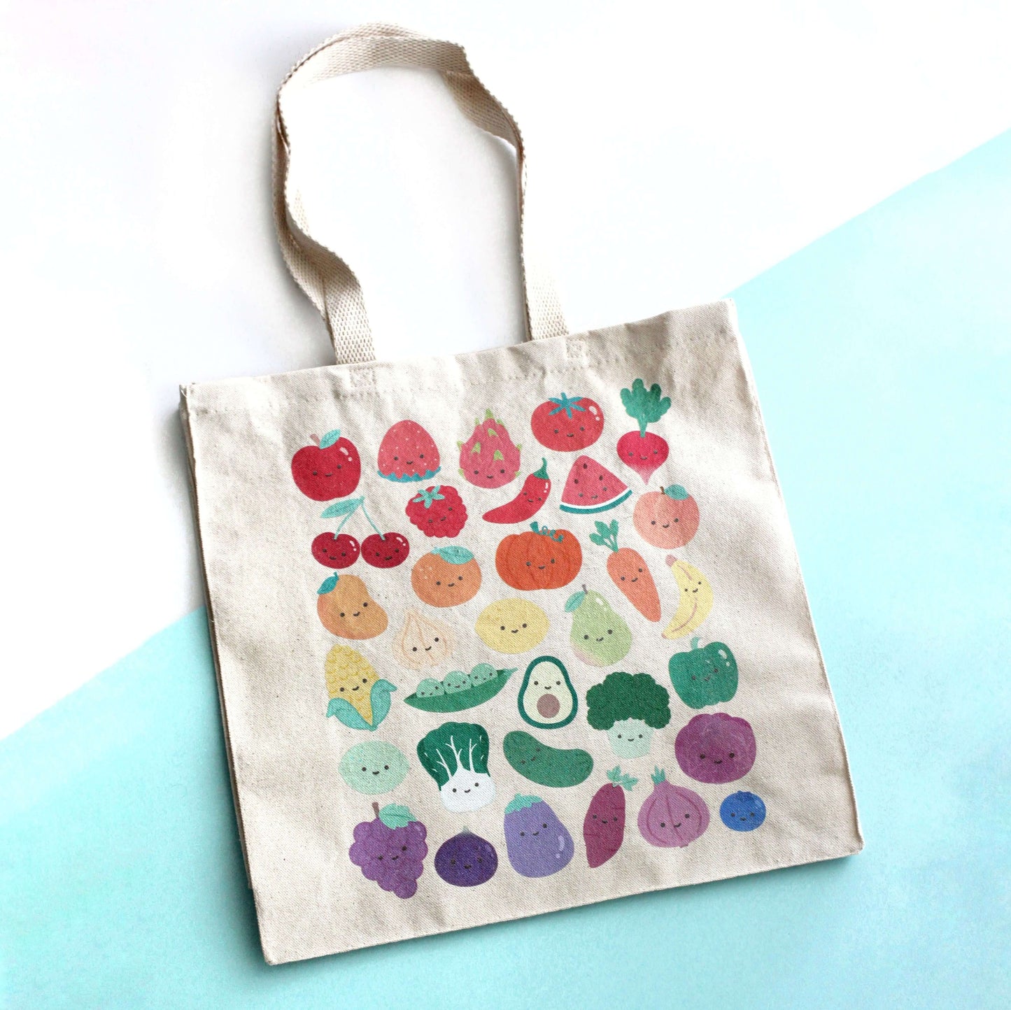 Tote bag with print of colorful fruits and vegetables with smiling faces on a light blue surface