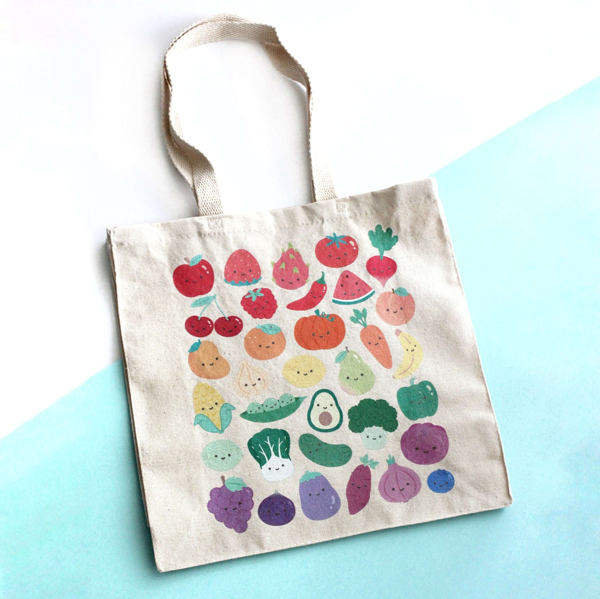 Tote bag with print of colorful fruits and vegetables with smiling faces on a light blue surface