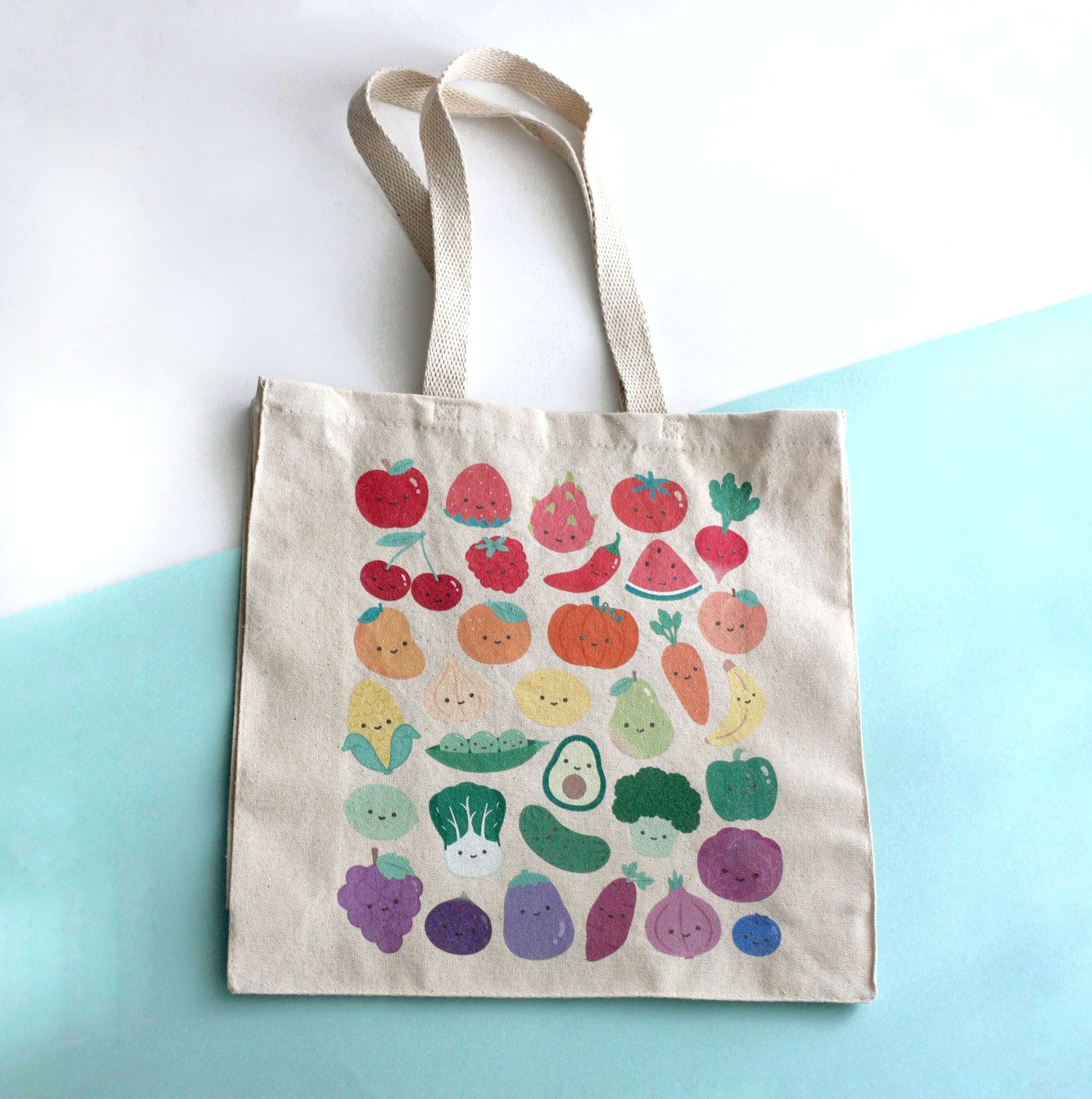 Tote bag with print of colorful fruits and vegetables with smiling faces on a light blue background. 