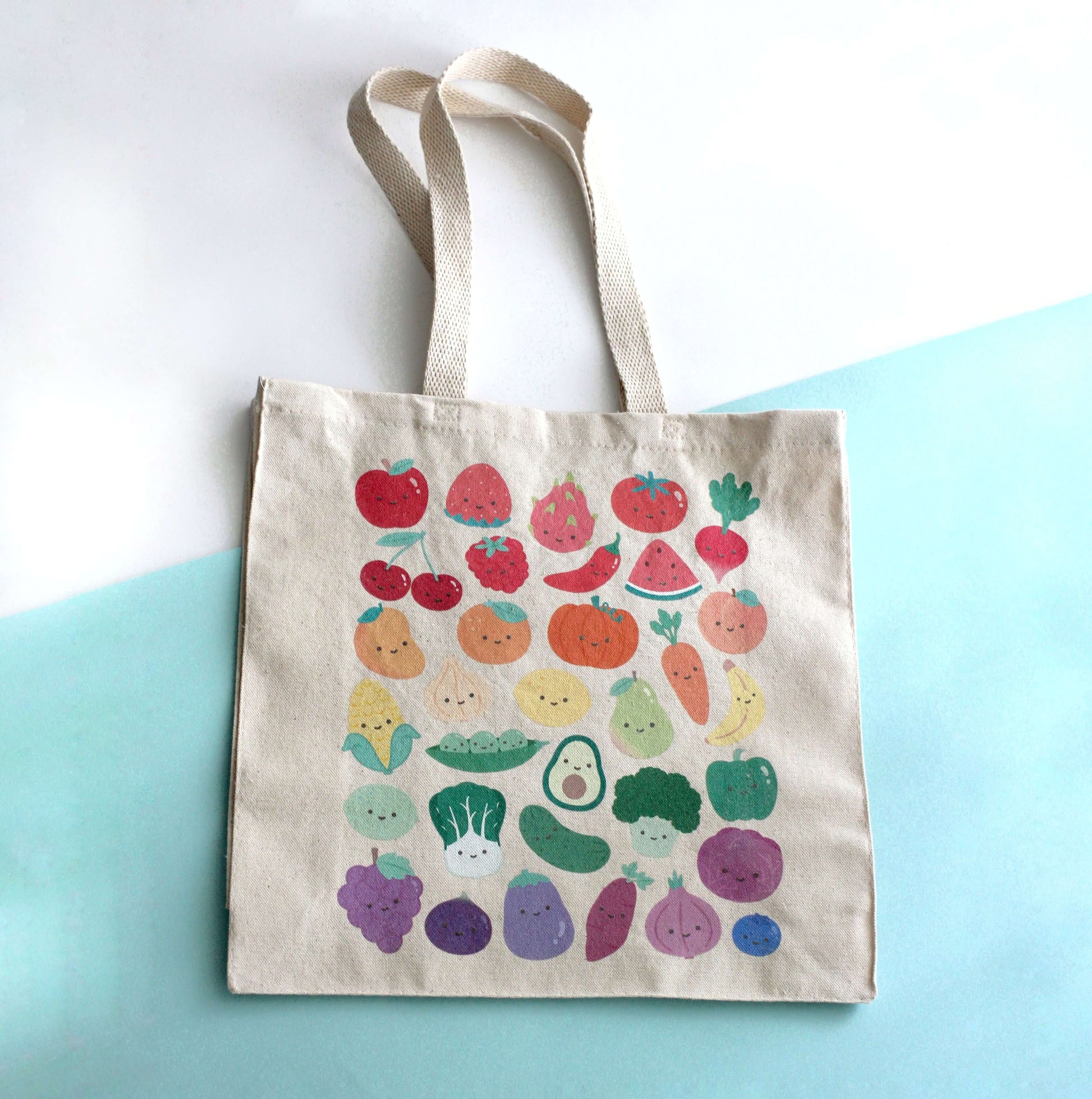 Tote bag with print of colorful fruits and vegetables with smiling faces on a light blue background. 
