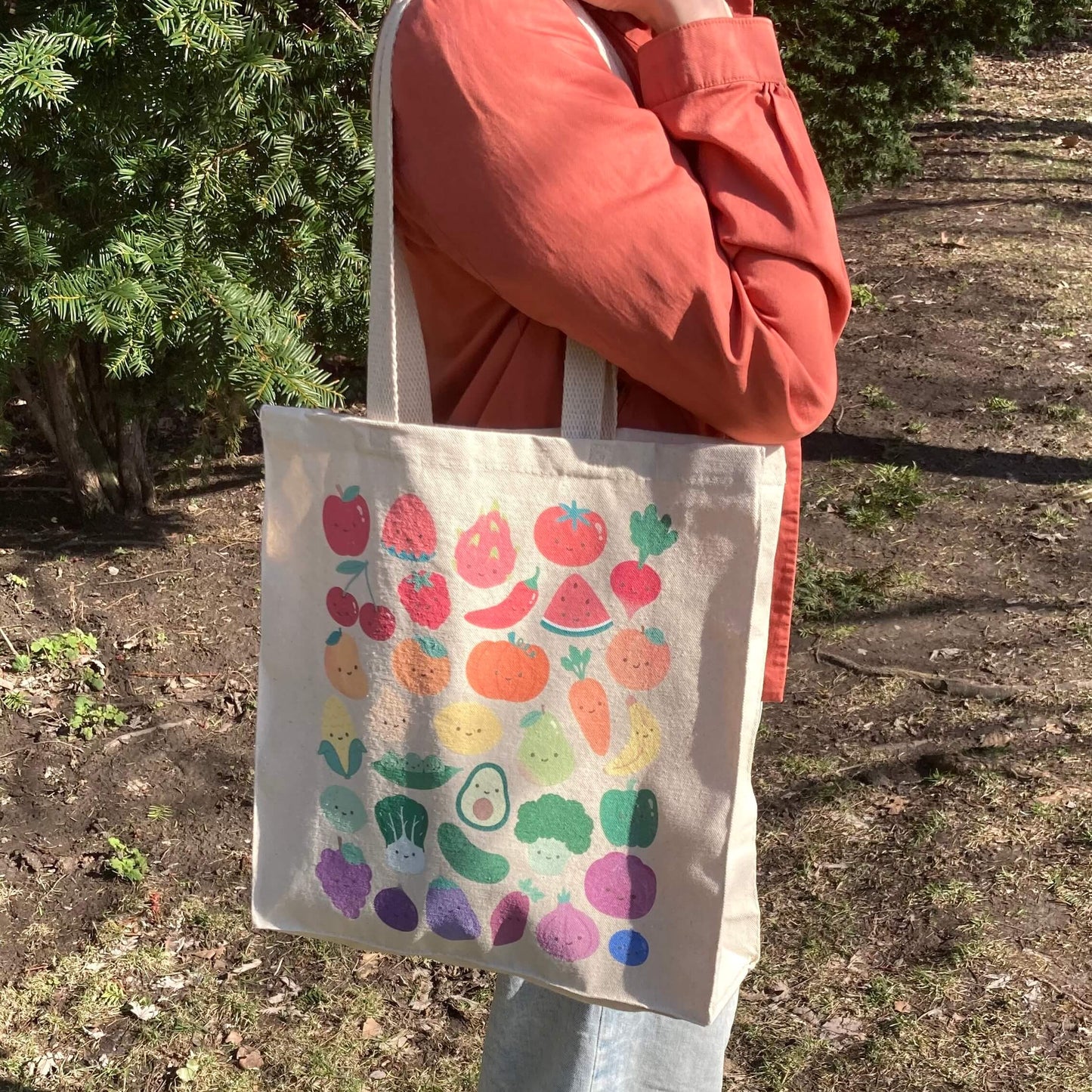 Tote bag with print of colorful fruits and vegetables with smiling faces held by a person outdoors.