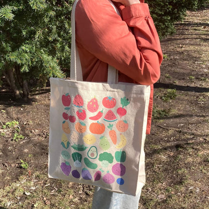 Tote bag with print of colorful fruits and vegetables with smiling faces held by a person outdoors.
