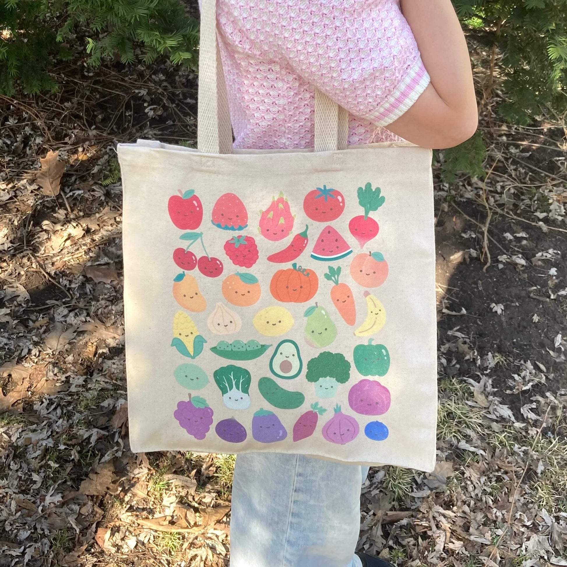 Tote bag with print of colorful fruits and vegetables with smiling faces held by a person outdoors.