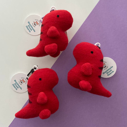 Three small plush dinosaur keychains in bright red with black stitched details and white circular tags, arranged on a split white and lavender background.